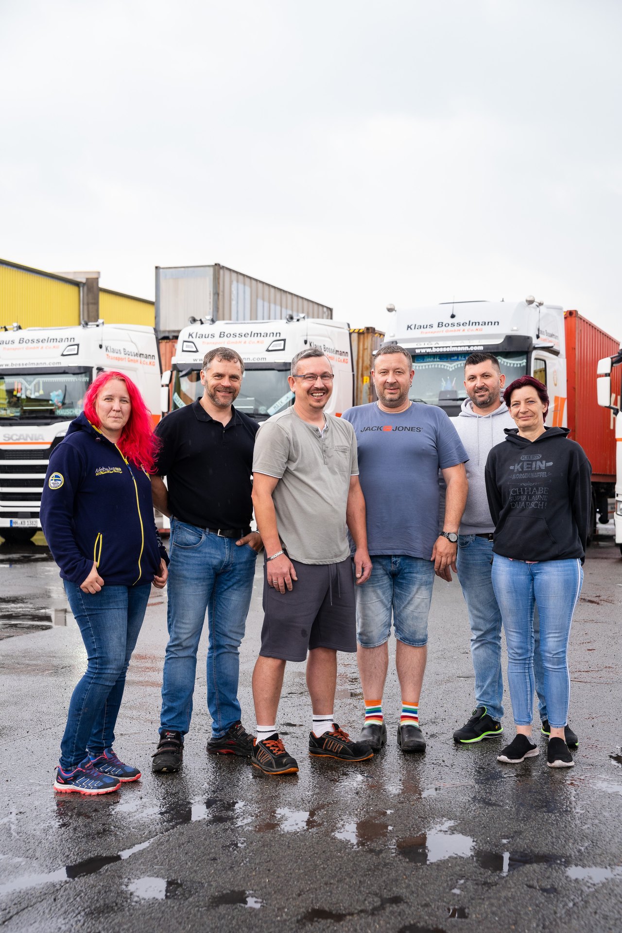 Sechs lächelnde Personen stehen vor weißen Lastwagen der Firma "Klaus Bosselmann" auf nassem Asphalt.